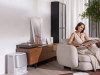 Eine Frau liest eine Zeitschrift auf einem Sofa, während ein Staubsauger-Roboter auf dem Boden steht.