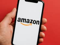 Eine Hand hält ein Smartphone, das das Amazon-Logo auf einem weißen Bildschirm vor rotem Hintergrund zeigt.