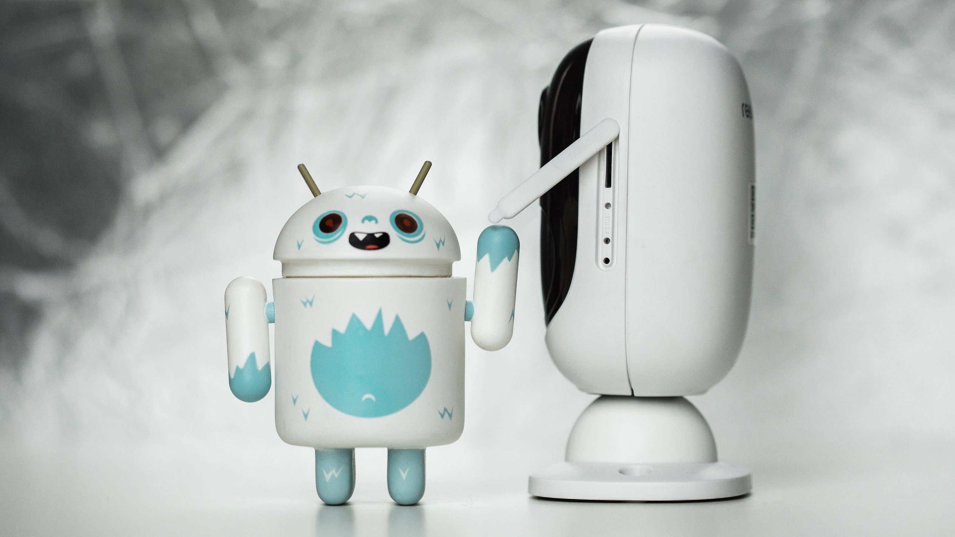 Ein niedlicher Spielzeugroboter mit blauen Akzenten steht neben einer weißen Reolink-Kamera.