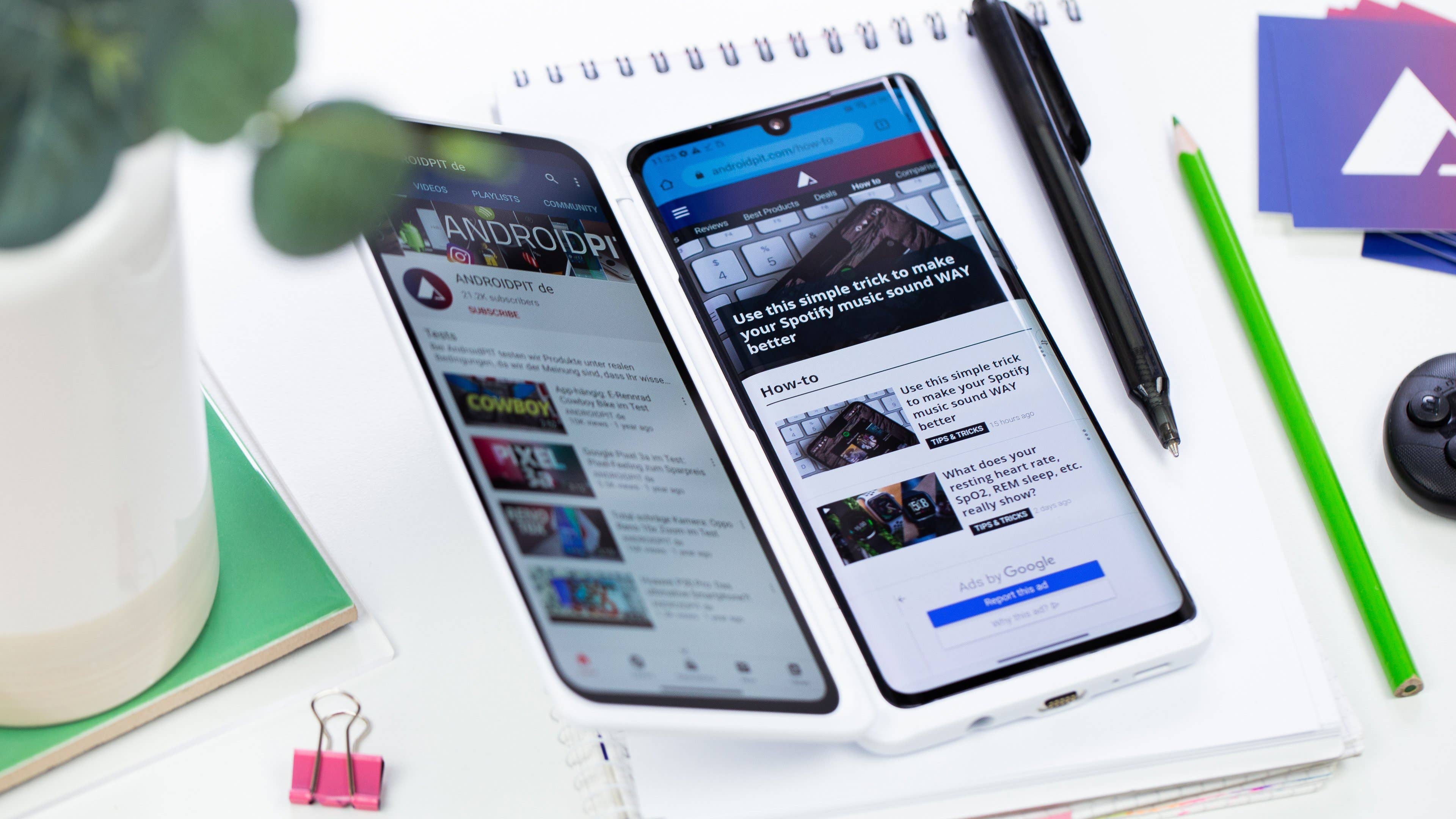 Zwei Smartphones, die Websites auf einem Tisch mit einem Notizbuch, einer Pflanze und Schreibwaren anzeigen.