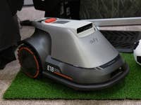 Eufy E18-Rasenmäher auf Kunstrasen mit dekorativen Pflanzen und einem Fußball in der Nähe.