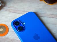 Ein blaues Smartphone neben einem orangefarbenen Apple AirTag und gelben Ohrhörern auf einer Holzoberfläche.