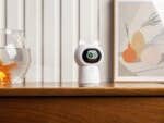 Aqara-Kamera-Hub G3 auf einem Holztisch neben einem Fischglas und einem gerahmten abstrakten Kunstwerk.