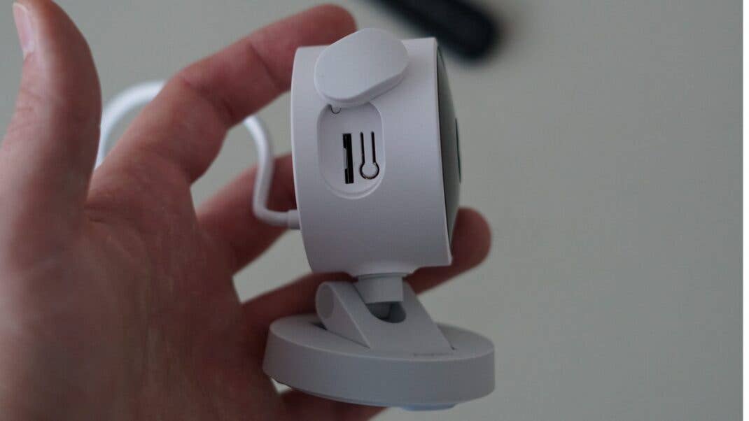 Eine Hand hält eine weiße Aqara G100 Kamera mit einem Kabel und einer Seitenansicht, die einen kleinen Schlitz zeigt. Eine Hand hält eine weiße Aqara G100 Kamera mit einem Kabel und einer Seitenansicht, die einen kleinen Schlitz zeigt.