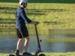 Person fährt einen Ausom L1 EU E-Scooter auf einem Weg am Wasser.