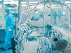 Ein transparenter menschlicher Kopfmodell mit Drähten und Sensoren in einem Labor, im Hintergrund eine Person in Schutzausrüstung.