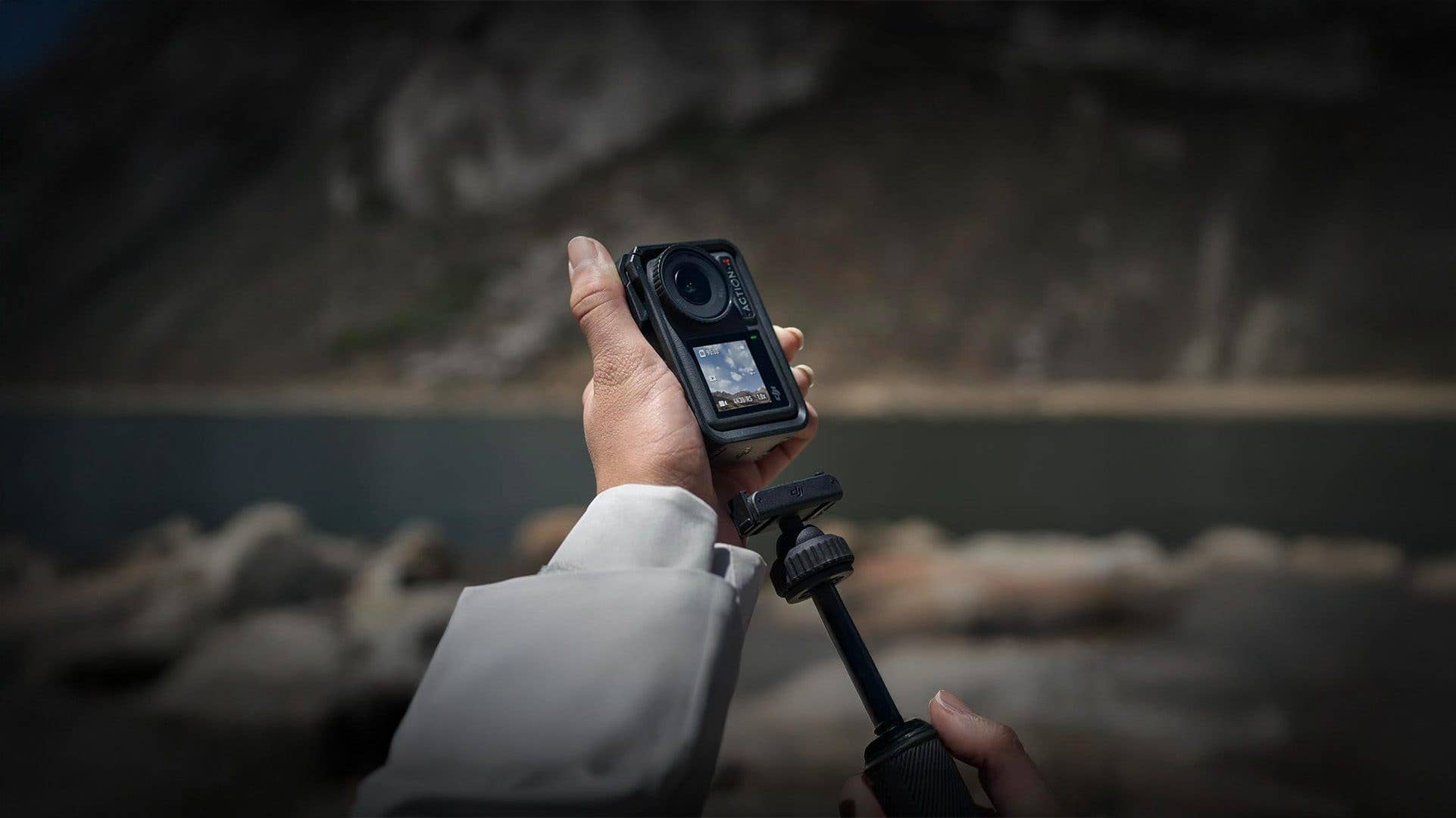 Eine Person hält eine DJI Osmo Action 4 Kamera auf einem Stativ in der Nähe eines Gewässers.