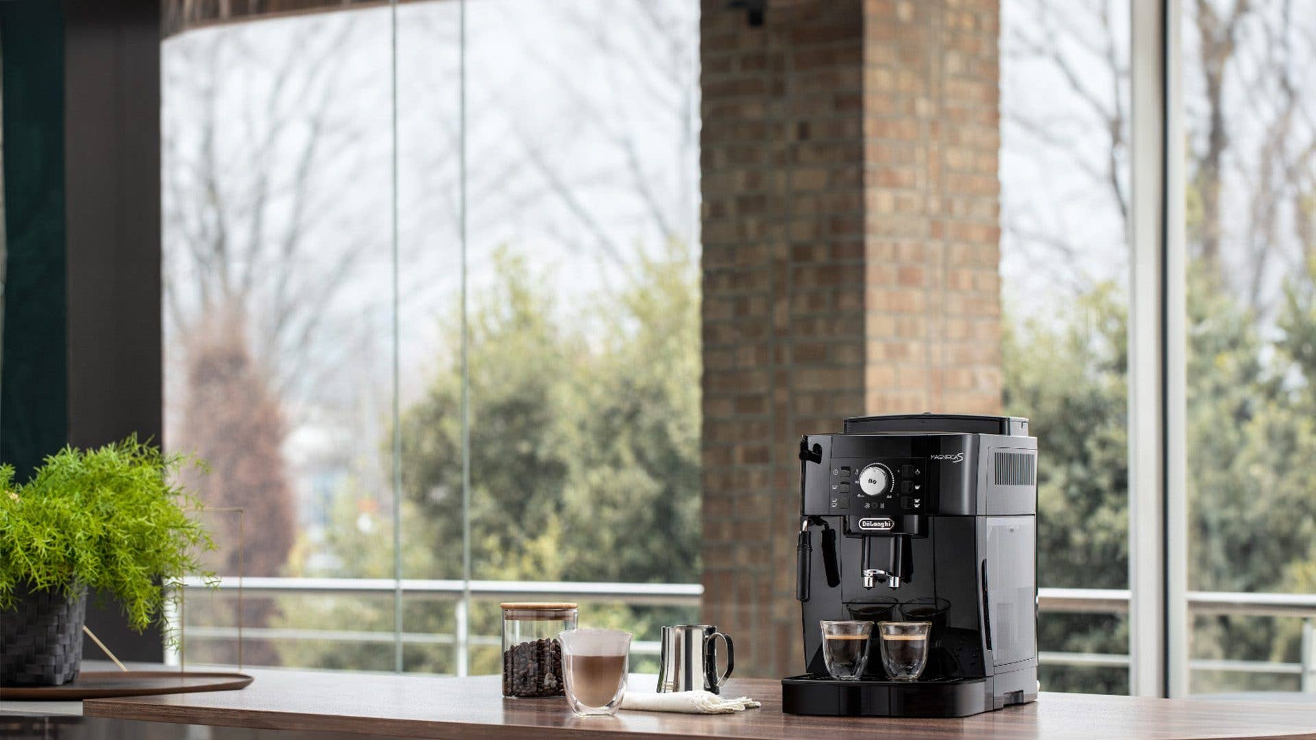 Eine De'Longhi Magnifica S Kaffeemaschine auf einem Holztisch mit Kaffeetassen und einer Pflanze in heller Umgebung.