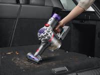 Eine Person benutzt einen Dyson V8 kabellosen Staubsauger, um den Kofferraum eines Autos mit Schmutz und Ablagerungen zu reinigen.