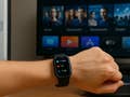 Eine Hand mit einer Smartwatch, die Herzfrequenz und Schritte anzeigt, im Hintergrund ein Fernseher mit verschiedenen Apps.