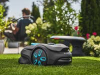 Ein Gardena Smart Sileno Sense Rasenmähroboter auf grünem Gras mit einer Person im Hintergrund, die sich entspannt.