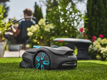 Ein Gardena Smart Sileno Sense Rasenmähroboter auf grünem Gras mit einer Person im Hintergrund, die sich entspannt.
