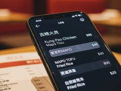 Ein Smartphone zeigt ein Menü mit Gerichten wie Kung Pao Chicken und Mapo Tofu, einschließlich Preisen in einem Restaurant.