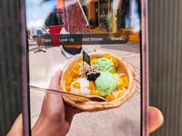 Eine Hand hält eine Schüssel mit buntem Eis in Mango-, grünen und weißen Geschmacksrichtungen, auf einem Smartphone-Bildschirm.