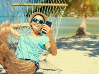 Eine Frau, die in einer Hängematte am Strand entspannt, ein Smartphone benutzt und Sonnenbrillen trägt.