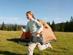 Ein Mädchen läuft im Freien und hält einen Jackery Explorer 500 v2 in der Nähe eines Zeltes auf einer Wiese.