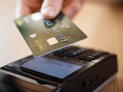 Eine Hand hält eine Kreditkarte über ein Kartenlesegerät auf einer Holzoberfläche.