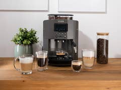 Eine Krups Essential EA8100 Kaffeemaschine mit verschiedenen Kaffeegetränken auf einem Holztisch.