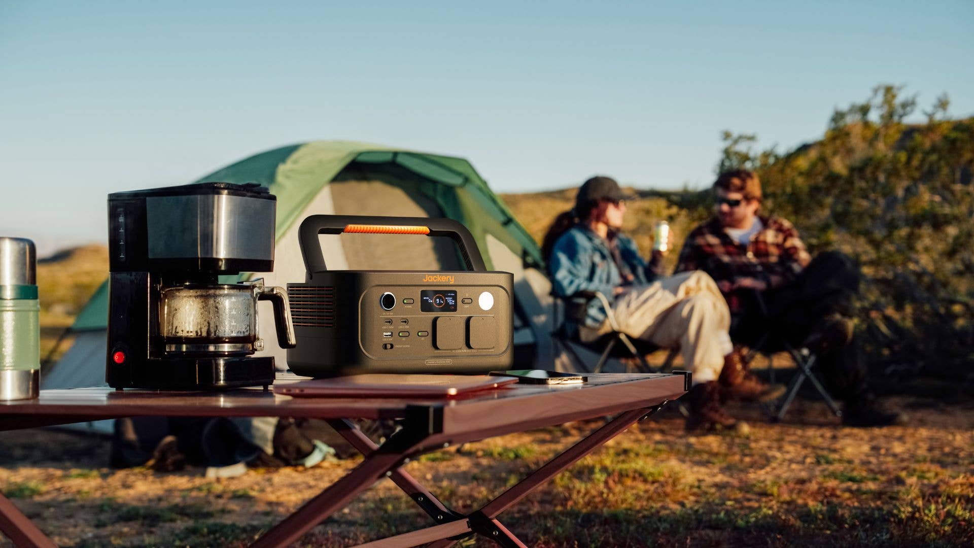 Eine Jackery-Stromstation auf einem Tisch, mit einer Kaffeemaschine und Menschen, die sich in der Nähe eines Zeltes entspannen.
