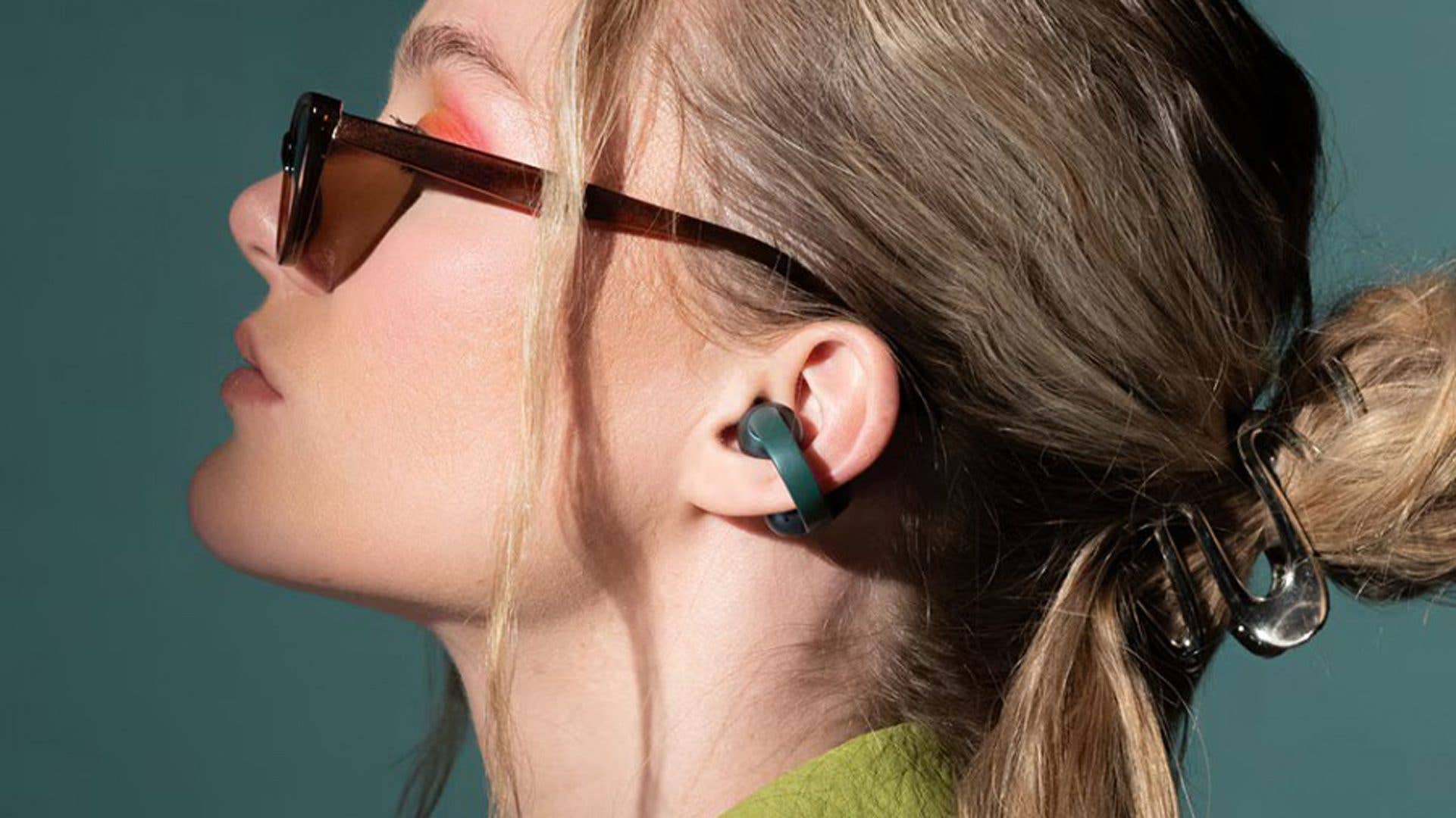 Eine Frau trägt Sonnenbrillen und Motorola Buds Loop-Ohrhörer, mit einem Pferdeschwanz und einem grünen Hintergrund.