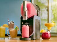 Ninja Slushmaschine mit verschiedenen bunten Getränken in Gläsern auf einem Holztisch.
