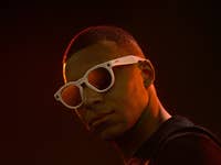 Eine Person mit weißen Sonnenbrillen vor einem dunklen Hintergrund.