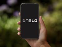 Eine Hand hält ein Smartphone, das das Otelo-Logo auf dunklem Hintergrund zeigt.