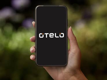 Eine Hand hält ein Smartphone, das das Otelo-Logo auf dunklem Hintergrund zeigt.