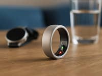 Nahaufnahme eines Oura-Smartrings auf einem Holztisch mit einem Glas Wasser und einer Smartwatch im Hintergrund.