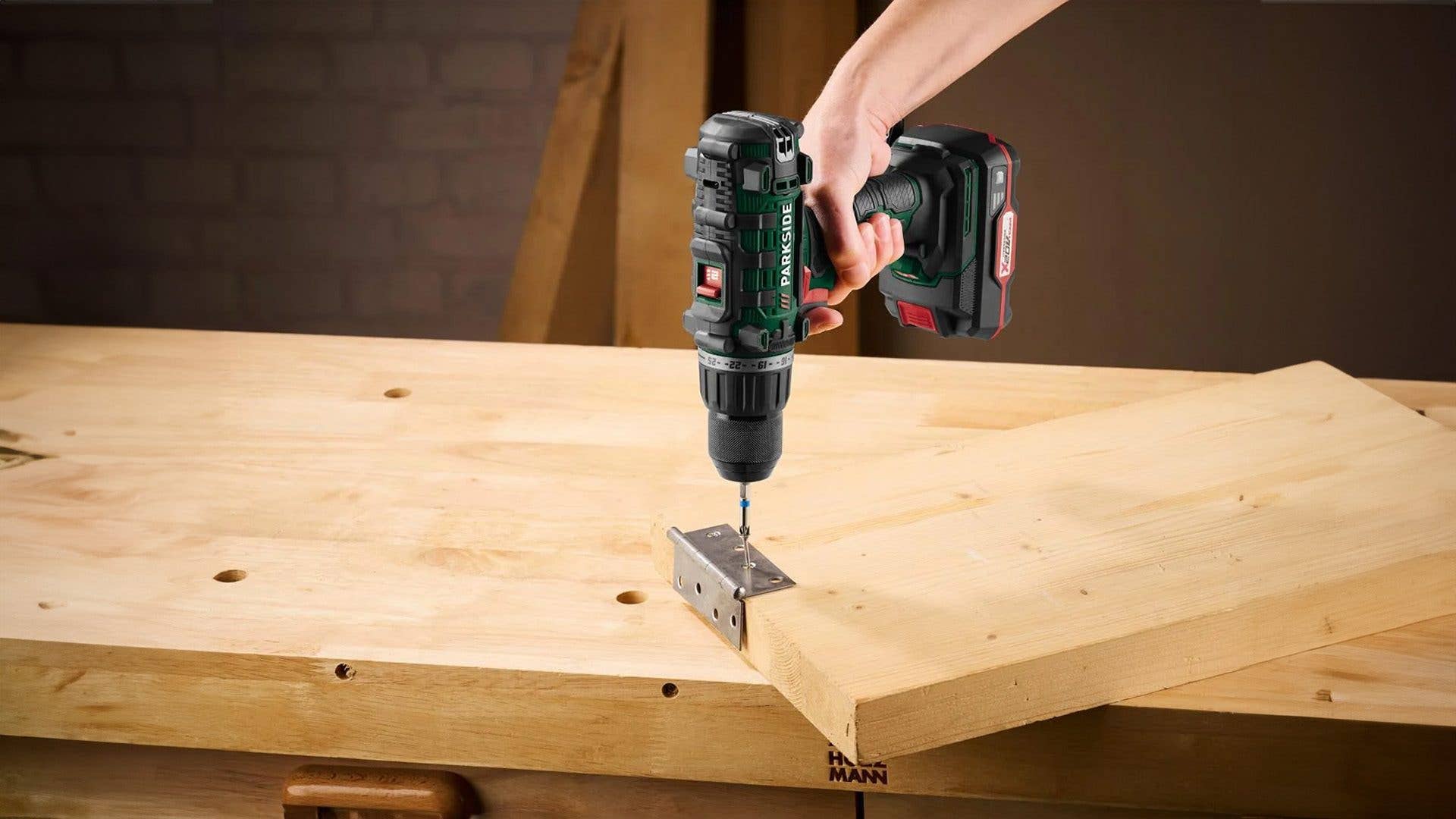 Eine Hand hält einen Parkside-Akkuschrauber auf einer Holzoberfläche, der in ein Metall-Scharnier bohrt.