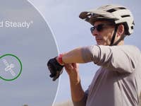 Ein Radfahrer mit Helm schaut auf eine Smartwatch, mit dem Text 'Hold Steady' und einem Satelliten-Symbol.