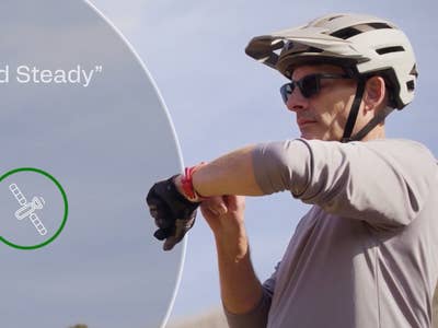 Ein Radfahrer mit Helm schaut auf eine Smartwatch, mit dem Text 'Hold Steady' und einem Satelliten-Symbol.