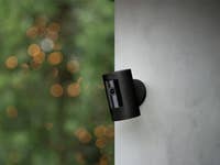 Eine schwarze Ring-Sicherheitskamera, die an einer Wand montiert ist, vor einem unscharfen grünen Hintergrund.