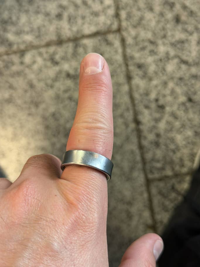 Eine Hand mit einem metallenen Samsung Galaxy Ring am Daumen, vor einer strukturierten Oberfläche.