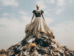 Eine Schaufensterpuppe steht in einem wallenden Rock oben auf einem Müllberg aus Wegwerf-Mode