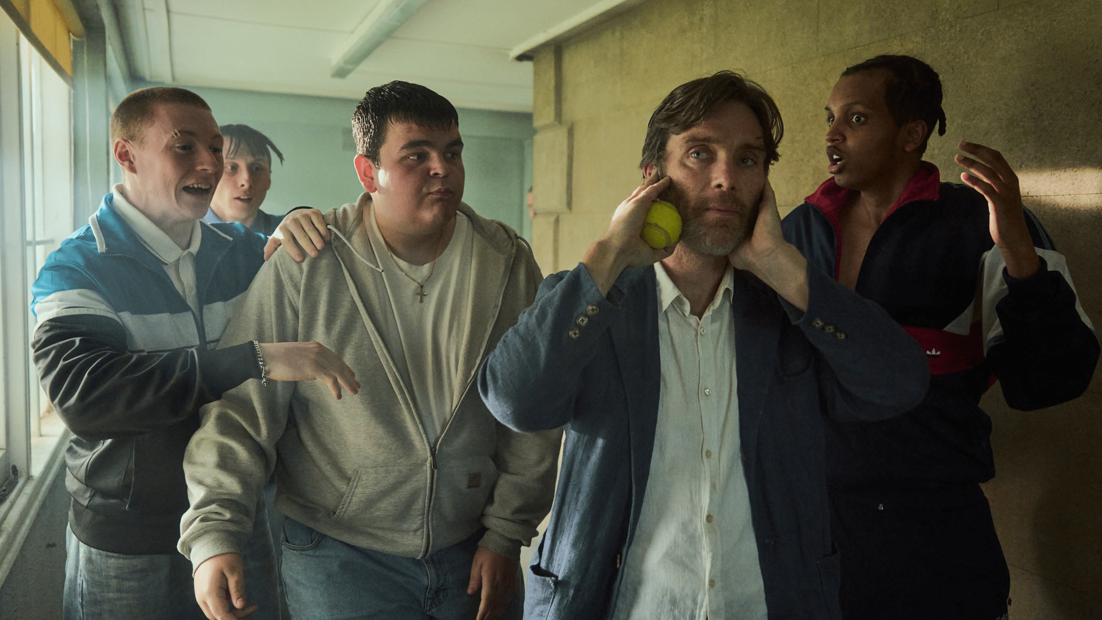 Eine Gruppe von vier jungen Männern umgibt einen besorgten Mann, der sich den Kopf hält, während einer einen Tennisball wirft.