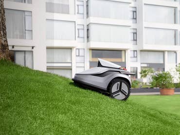 Ein Mähroboter auf einer geneigten Grasfläche neben einem Baum und modernen Gebäuden im Hintergrund.