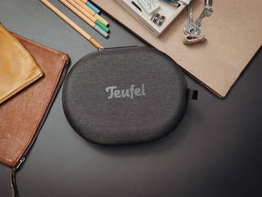 Eine graue Kopfhörertasche mit 'Teufel'-Logo auf einem Tisch, umgeben von Schreibwaren.