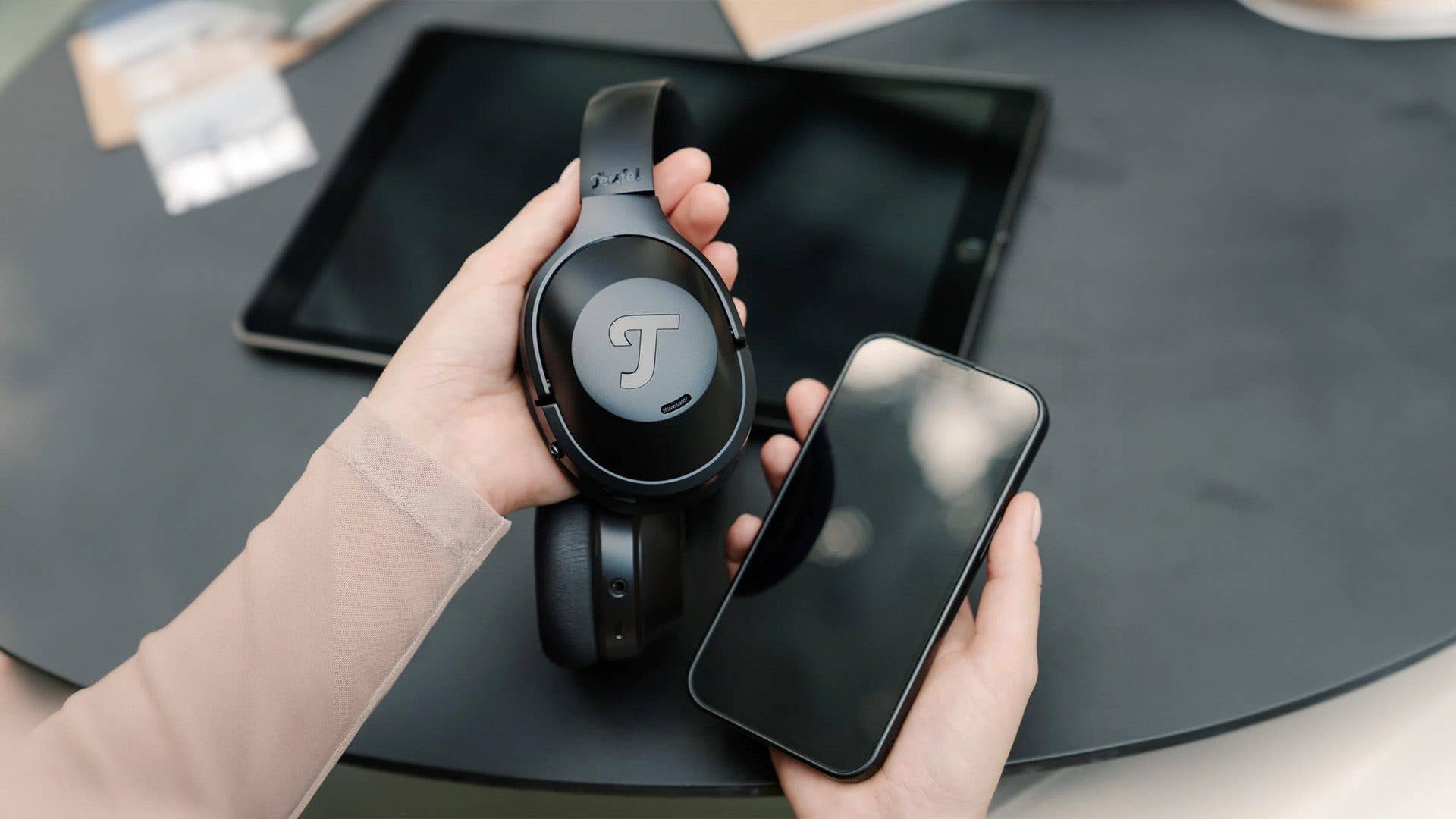 Eine Person hält schwarze Teufel Over-Ear-Kopfhörer und ein Smartphone auf einem Tisch.