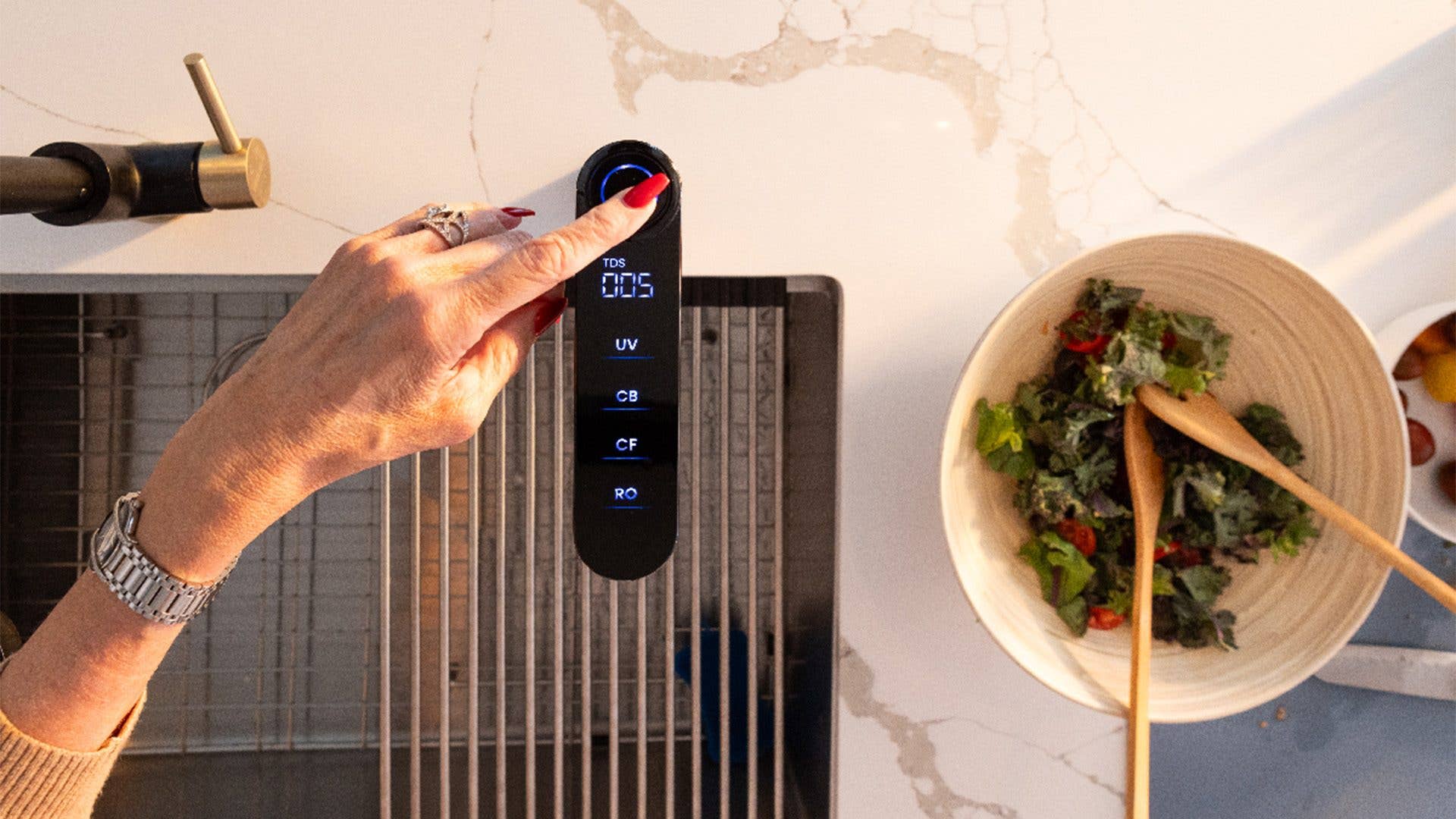 Eine Hand mit roten Nägeln drückt einen Knopf an einem Wasserqualitätsmonitor neben einer Schüssel Salat.