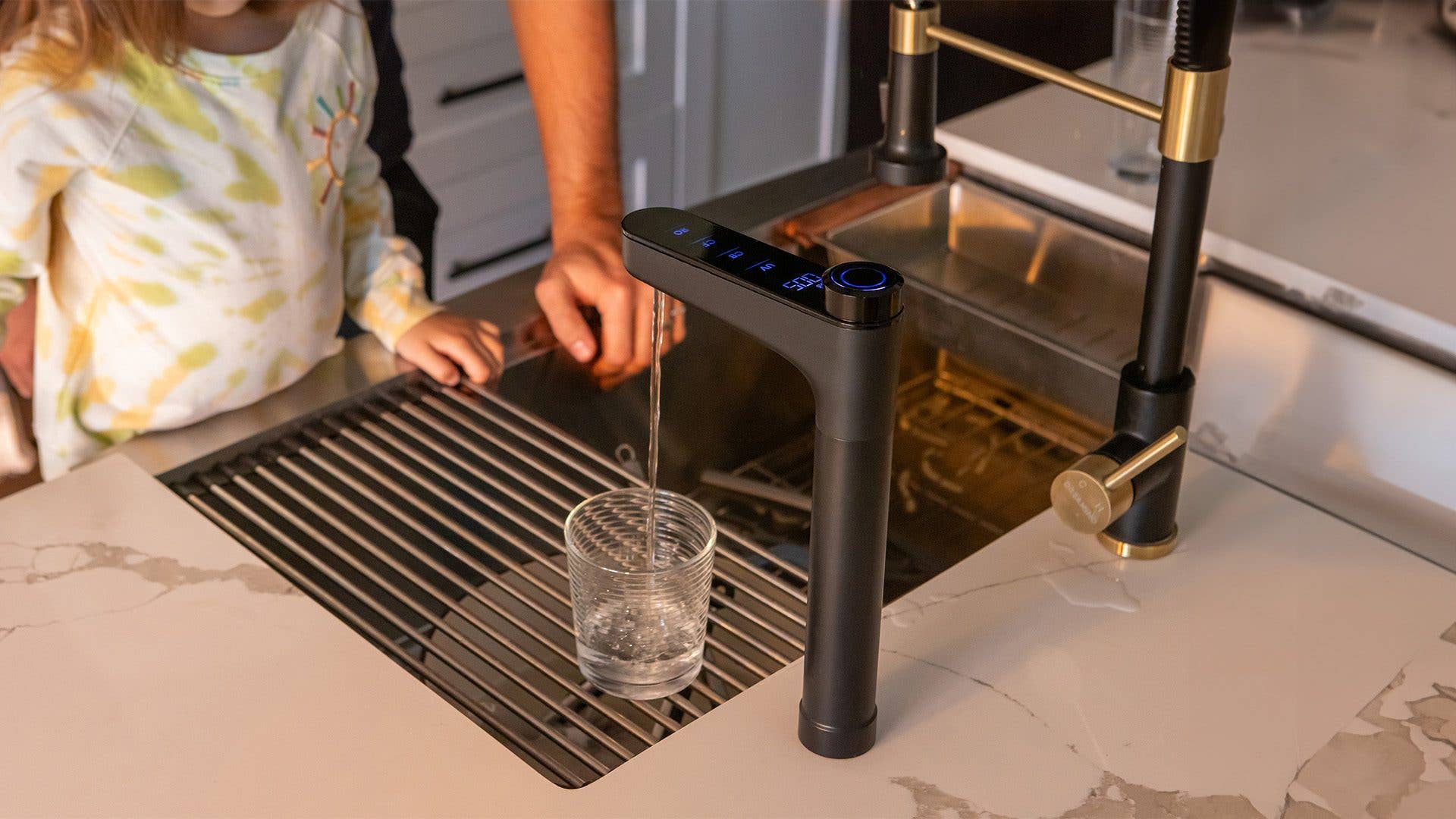 Ein Kind schaut zu, wie Wasser aus einem modernen Wasserhahn in ein Glas auf einer Küchenarbeitsplatte fließt.