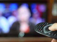 Eine Hand hält eine Fernbedienung mit einem verschwommenen Fernseher im Hintergrund, auf dem eine Person zu sehen ist.