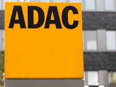 Ein großes gelbes Schild mit dem Text 'ADAC' vor einem verschwommenen Hintergrund von Fensterfassaden.