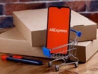 AliExpress-Logo auf einem Smartphone, das in einem kleinen Einkaufswagen vor einigen paketen steht.