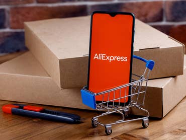 AliExpress-Logo auf einem Smartphone, das in einem kleinen Einkaufswagen vor einigen paketen steht.