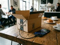 Amazon Paket aus dem ein Kabel herausschaut steht in einem Kaffee und ist mit einem Apple iPhone 17 Pro Max verbunden.