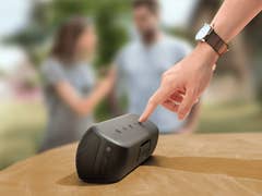 Eine Hand, die auf den Anker Soundcore Motion Plus Lautsprecher auf einem Tisch zeigt, mit zwei unscharfen Personen im Hintergrund.