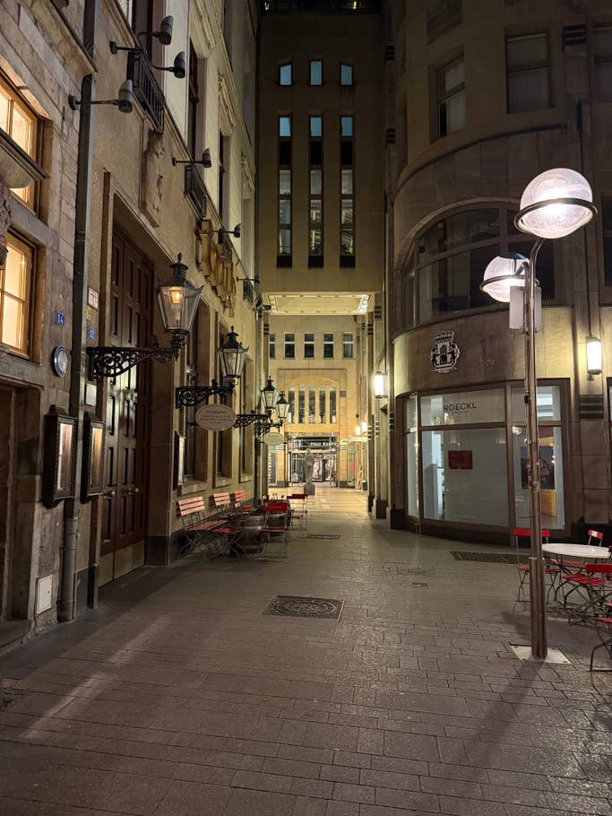 Eine enge Gasse bei Nacht mit beleuchteten Straßenlaternen, leeren Stühlen und historischen Gebäuden.