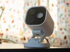 Eine weiße Aqara-Kamerahub G5 Pro auf einer schwarzen Oberfläche mit einem bunten gemusterten Hintergrund.