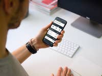 Eine Person nutzt die comdirect-App auf einem Smartphone an einem Schreibtisch mit Computer und Tastatur.